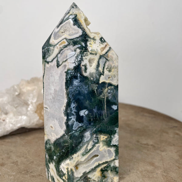 Moss Agate Tower 14cm standing on stone bench in front of crystal cluster